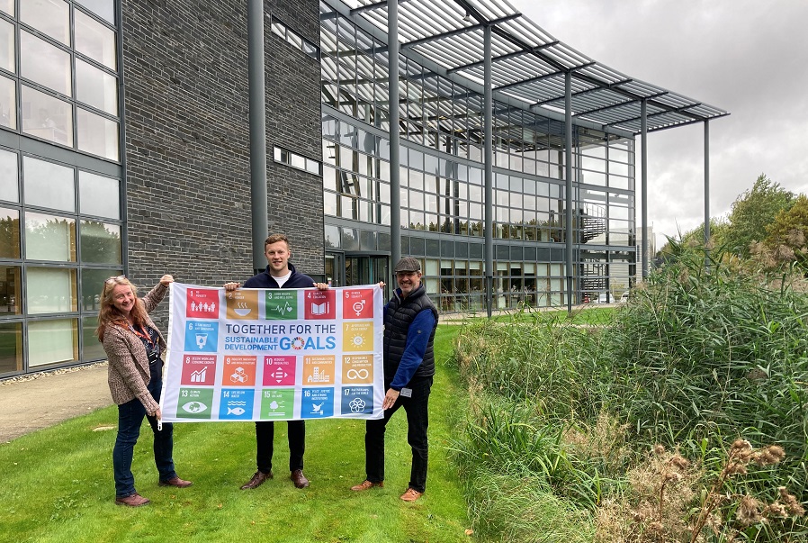Three people holding SDG flag in front of office building in Howbery Park, Oxfordshire