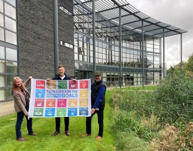 Three people holding SDG flag in front of office building in Howbery Park, Oxfordshire