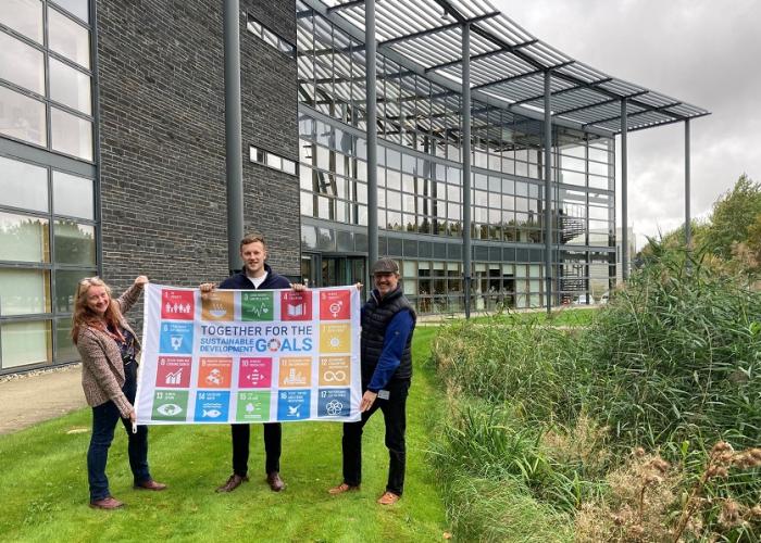 Three people holding SDG flag in front of office building in Howbery Park, Oxfordshire