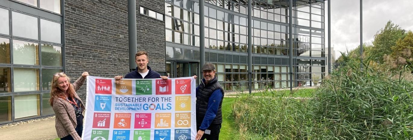 Three people holding SDG flag in front of office building in Howbery Park, Oxfordshire